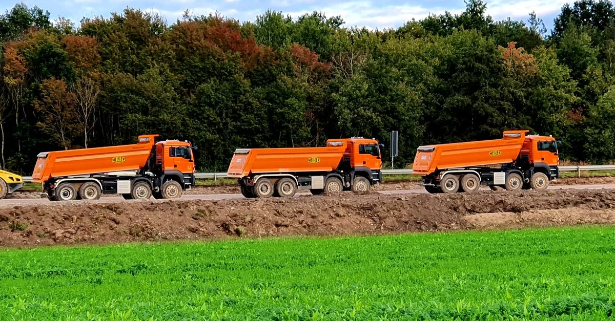 Trois camions-bennes oranges garés sur un site de construction en plein air à côté de champs de gazon vert.