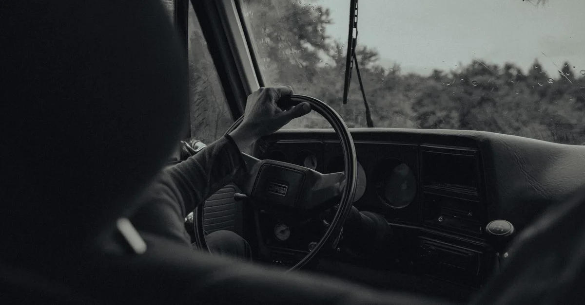 Une vue en noir et blanc d'une personne conduisant une voiture vintage, mettant en valeur le volant et le tableau de bord.
