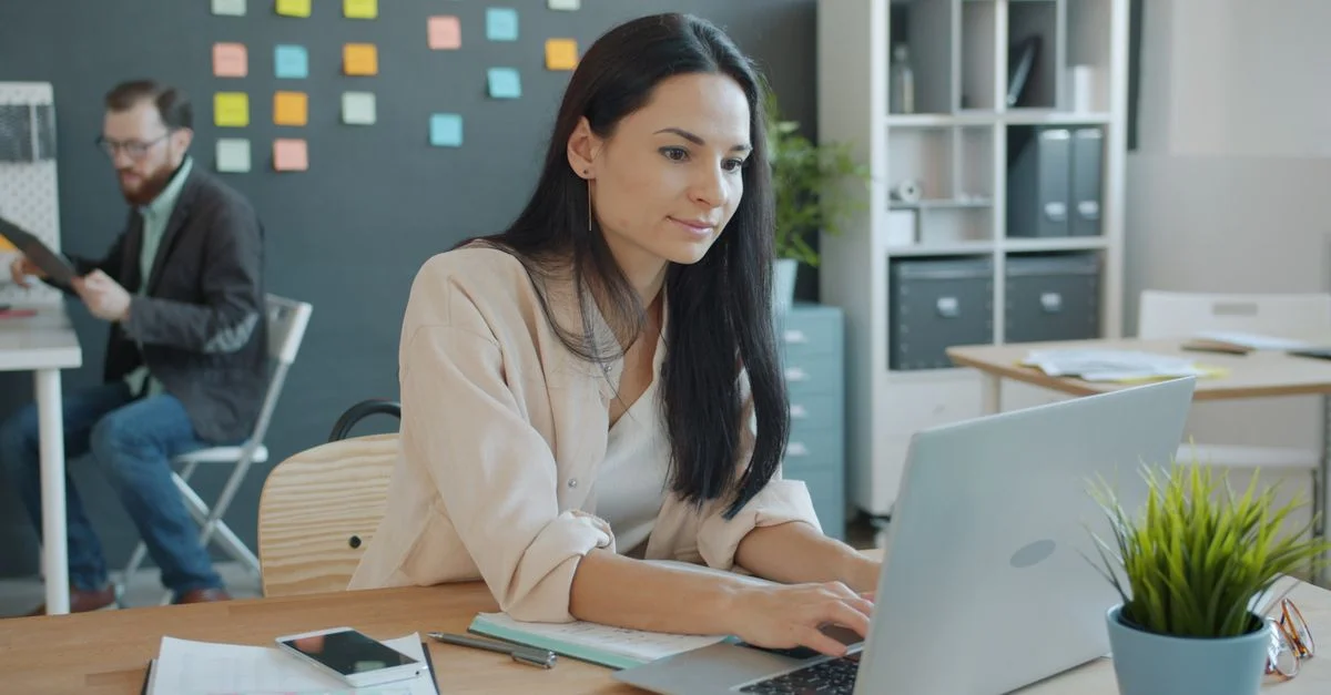 Femme travaillant sur un ordinateur portable dans un bureau moderne, avec une collègue en arrière-plan.