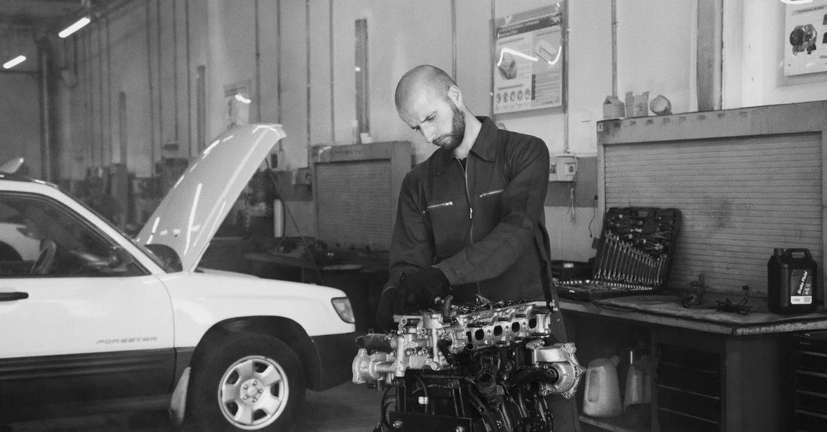 Photo monochrome d'un mécanicien inspectant un moteur de voiture dans un garage. Outils visibles en arrière-plan.