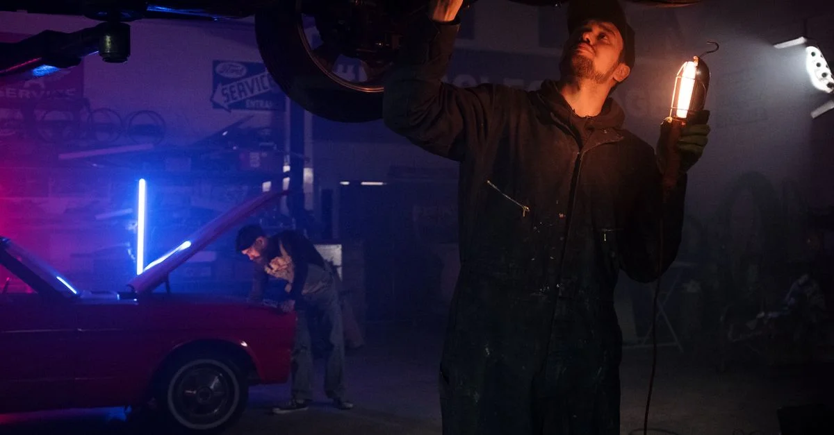 Mécanicien travaillant sur un véhicule sous des lumières néon dans un garage sombre, mettant en avant ses compétences en réparation automobile.